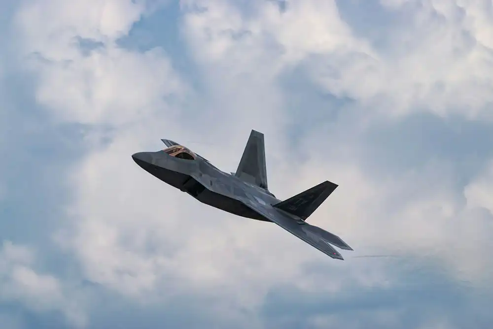 A U.S. Air Force F-22 Raptor fighter jet flying at an upward angle against a backdrop of partly cloudy sky.